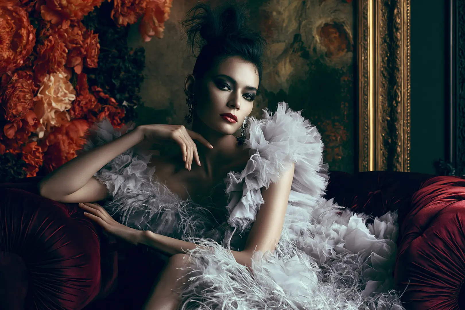 Fashion-Editorial einer Frau im Federkleid auf einem Samtsofa umgeben von Blumen und einem Ölgemälde. Dramatische Lichtstimmung .
